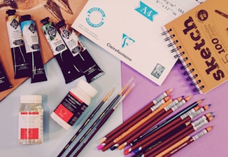 A variety of art supplies are spread out, including tubes of paint, jars of medium, colored pencils, a sketchbook, and sheets of paper. There are several artist paint brushes alongside pencils of different colors arranged neatly, with some paint tubes and medium bottles placed nearby.