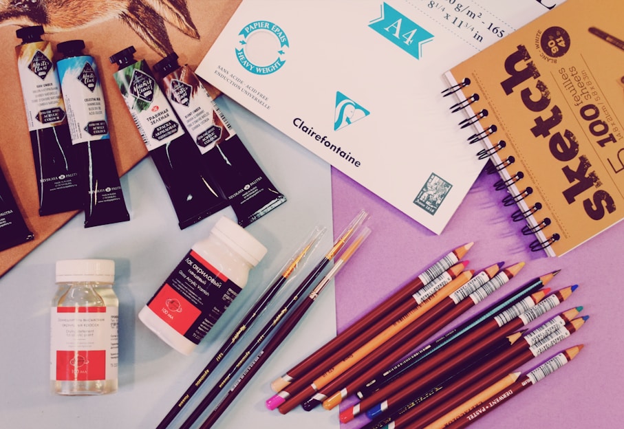 A variety of art supplies are spread out, including tubes of paint, jars of medium, colored pencils, a sketchbook, and sheets of paper. There are several artist paint brushes alongside pencils of different colors arranged neatly, with some paint tubes and medium bottles placed nearby.