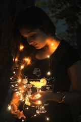 A person is gently holding a string of glowing fairy lights against a dark background. The warm, orange light softly illuminates their face and hands, creating a serene and intimate atmosphere. The person is wearing a dark shirt with light patterns and illustrations on it.