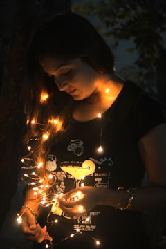 A person is gently holding a string of glowing fairy lights against a dark background. The warm, orange light softly illuminates their face and hands, creating a serene and intimate atmosphere. The person is wearing a dark shirt with light patterns and illustrations on it.