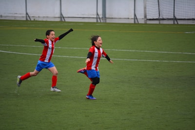 Two children wearing red and white striped jerseys and blue shorts are joyfully running on a soccer field. Both appear to be celebrating with their arms outstretched, expressing excitement and happiness.