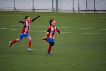Two children wearing red and white striped jerseys and blue shorts are joyfully running on a soccer field. Both appear to be celebrating with their arms outstretched, expressing excitement and happiness.