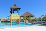 A water park featuring a large water slide structure and a building with a brown roof. The slide has a blue and red color scheme. There is a signboard at the base of the slide and various aquatic features including a small fountain in the foreground. Surrounding the area are palm trees and well-maintained landscaping. The sky is clear and bright blue.
