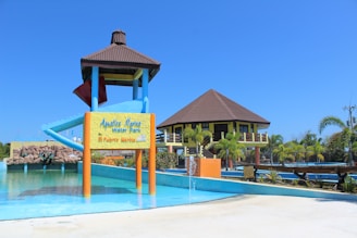 A water park featuring a large water slide structure and a building with a brown roof. The slide has a blue and red color scheme. There is a signboard at the base of the slide and various aquatic features including a small fountain in the foreground. Surrounding the area are palm trees and well-maintained landscaping. The sky is clear and bright blue.