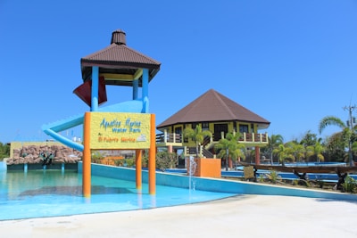 A water park featuring a large water slide structure and a building with a brown roof. The slide has a blue and red color scheme. There is a signboard at the base of the slide and various aquatic features including a small fountain in the foreground. Surrounding the area are palm trees and well-maintained landscaping. The sky is clear and bright blue.