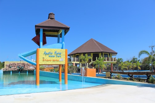 A water park featuring a large water slide structure and a building with a brown roof. The slide has a blue and red color scheme. There is a signboard at the base of the slide and various aquatic features including a small fountain in the foreground. Surrounding the area are palm trees and well-maintained landscaping. The sky is clear and bright blue.