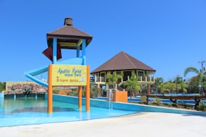 A water park featuring a large water slide structure and a building with a brown roof. The slide has a blue and red color scheme. There is a signboard at the base of the slide and various aquatic features including a small fountain in the foreground. Surrounding the area are palm trees and well-maintained landscaping. The sky is clear and bright blue.