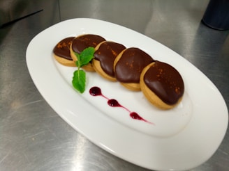 A plate with five round dessert cookies topped with a layer of glossy chocolate and a sprinkle of orange zest, arranged in a row. A small sprig of fresh mint and a red berry sauce accompany the cookies for garnish. The plate is positioned on a shiny metallic surface, likely a kitchen counter.