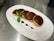 A plate with five round dessert cookies topped with a layer of glossy chocolate and a sprinkle of orange zest, arranged in a row. A small sprig of fresh mint and a red berry sauce accompany the cookies for garnish. The plate is positioned on a shiny metallic surface, likely a kitchen counter.