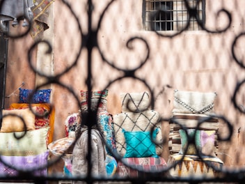 A collection of colorful and patterned cushions arranged against a textured wall, viewed through an artistic wrought iron fence. The cushions display a variety of designs, including stripes, checks, and geometric shapes, adding vibrancy to the background.