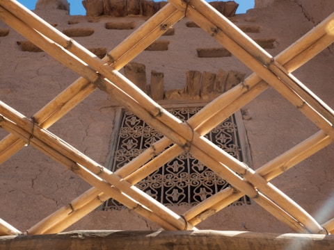 Woven, diagonal bamboo sticks create a grid in the foreground, framing a decorative, intricately patterned window. The background is a clay or adobe wall with windows that have rough wooden shutters.