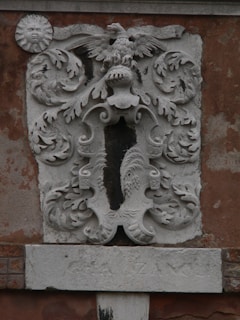 A detailed stone carving is displayed on a weathered wall, featuring an ornate coat of arms with a central eagle at the top and intricate patterns surrounding it. The design includes swirling acanthus leaves and a sun with a face on the left side of the carving. Beneath the coat of arms, there is an inscription partially etched into a stone slab.