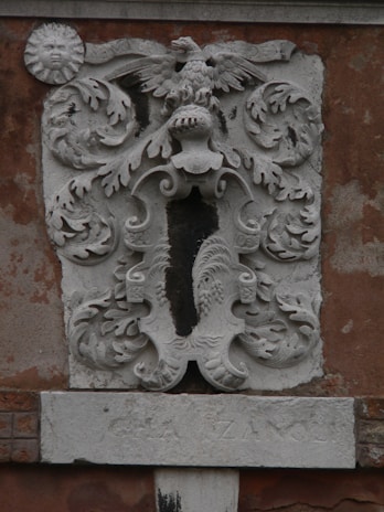 A detailed stone carving is displayed on a weathered wall, featuring an ornate coat of arms with a central eagle at the top and intricate patterns surrounding it. The design includes swirling acanthus leaves and a sun with a face on the left side of the carving. Beneath the coat of arms, there is an inscription partially etched into a stone slab.