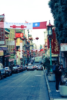 A vibrant street scene in a Chinatown district featuring numerous red lanterns hanging across the street. Buildings are adorned with traditional Chinese architectural features and signage, while flags are displayed overhead. The busy street is lined with parked cars and pedestrians walking along the sidewalks, contributing to an energetic and bustling atmosphere.