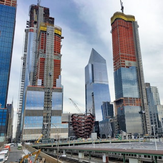 Skyscrapers under construction dominate the skyline, featuring cranes and scaffolding. A modern, honeycomb-like structure is visible in the center. The buildings are surrounded by an urban environment with a train station in the foreground.
