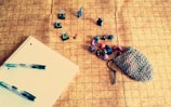 A tabletop gaming setup featuring a grid-patterned playmat. On the left, a lined notebook with two pens rests, while in the center, several painted miniature figurines are displayed. To the right, a chainmail pouch is partly open, spilling out colorful polyhedral dice of various shapes and numbers.
