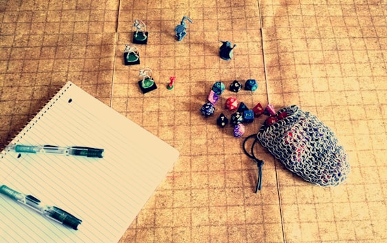 A tabletop gaming setup featuring a grid-patterned playmat. On the left, a lined notebook with two pens rests, while in the center, several painted miniature figurines are displayed. To the right, a chainmail pouch is partly open, spilling out colorful polyhedral dice of various shapes and numbers.