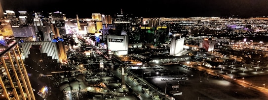 A panoramic view of a brightly lit city skyline at night, featuring numerous illuminated high-rise buildings and hotels. The scene captures a bustling urban atmosphere with vibrant lights casting reflections and creating a lively, energetic ambiance.