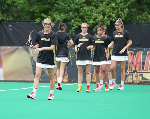 A group of female lacrosse players wearing black and gold Maryland jerseys and white shorts are standing together on a green field. They hold lacrosse sticks and are wearing protective eyewear. The background includes a fence with some foliage visible.