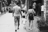 A black and white photo captures a family of three walking down a cobblestone street. The parents flank a small child as they walk, holding hands. The scene includes other pedestrians, a parked vehicle, and a clothing store displaying mannequins in the background. The atmosphere feels casual and unhurried.