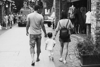 A black and white photo captures a family of three walking down a cobblestone street. The parents flank a small child as they walk, holding hands. The scene includes other pedestrians, a parked vehicle, and a clothing store displaying mannequins in the background. The atmosphere feels casual and unhurried.
