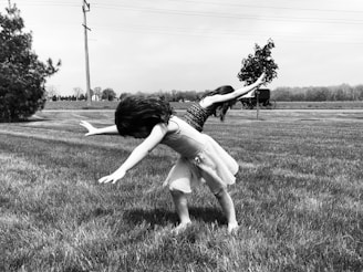 Two children are playing in a grassy field. They are captured in a dynamic pose, bending forward with their arms extended as if pretending to fly. The background features a rural landscape with a tree, power lines, and a horse-drawn carriage in the distance under a cloudy sky.