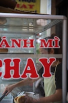 A person wearing an apron prepares a sandwich inside a small food stall. The words 'BÁNH MÌ CHAY' are prominently displayed on the front glass panel of the stall. The interior has ingredients and utensils visible through the glass.