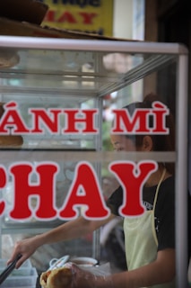 A person wearing an apron prepares a sandwich inside a small food stall. The words 'BÁNH MÌ CHAY' are prominently displayed on the front glass panel of the stall. The interior has ingredients and utensils visible through the glass.