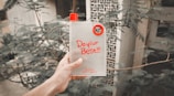 A hand is holding a transparent water bottle with a red cap and a circular red handle. The bottle features motivational text that reads 'Do your Best!!!' in bold red letters. In the background, there are blurred green leaves and part of a building with small square lattice openings.