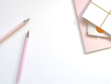 Two pink pens with metallic tips are placed diagonally on a light surface. Nearby, there are stacks of pastel-colored and patterned notebooks or cards.