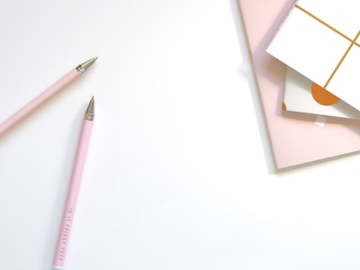 Two pink pens with metallic tips are placed diagonally on a light surface. Nearby, there are stacks of pastel-colored and patterned notebooks or cards.