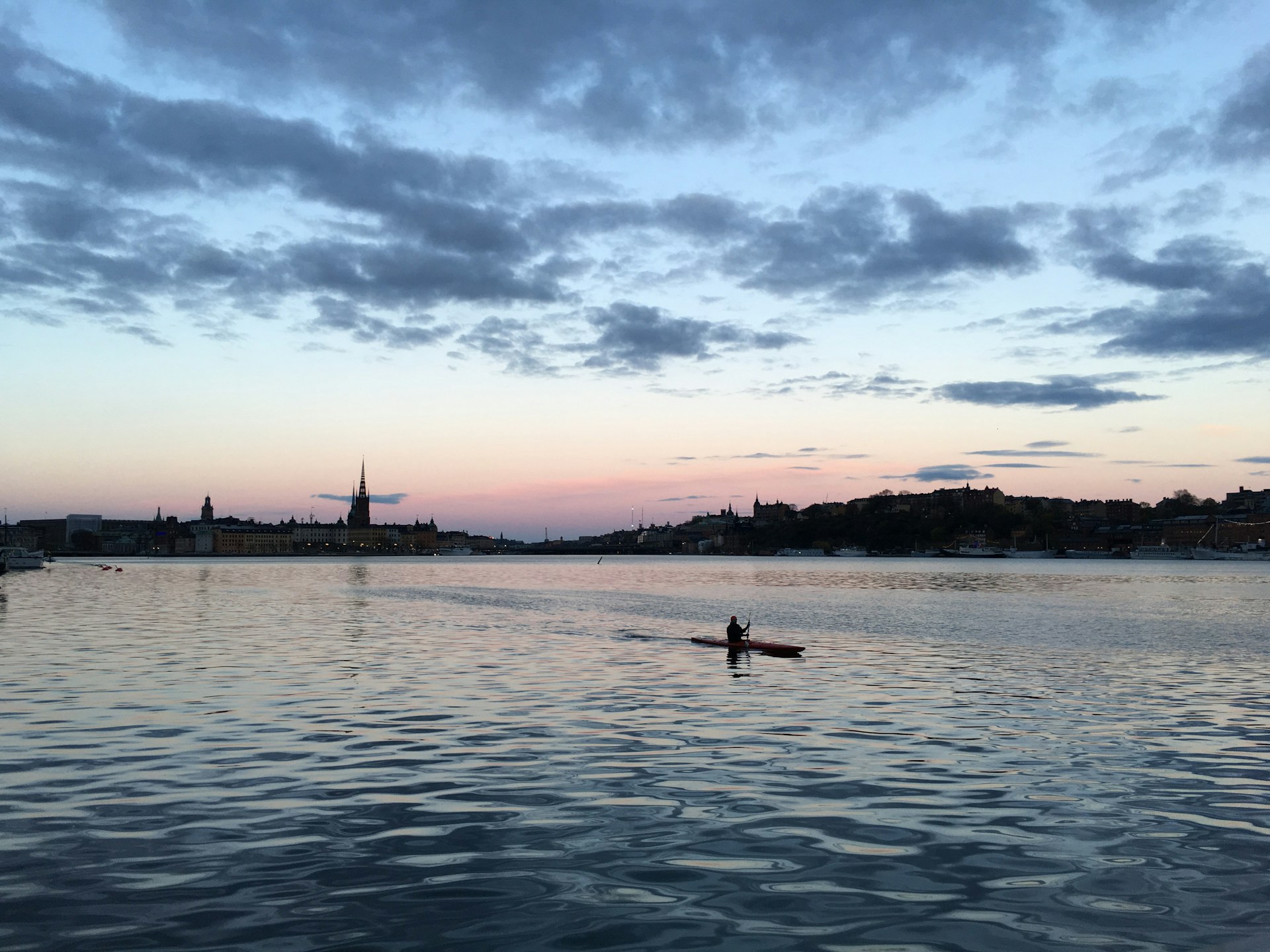 A kayaker paddles on a calm body of water during a serene sunset. The horizon features a silhouette of a cityscape with recognizable spires and historic buildings. Soft clouds hover in a sky transitioning from blue to pastel pink and orange hues.