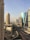 A cityscape featuring a row of tall modern buildings with glass facades. The architecture includes striking designs and a towering structure with a distinctive dome. The road below is lined with palm trees and appears empty with few cars. The sky is clear and slightly hazy.