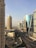 A cityscape featuring a row of tall modern buildings with glass facades. The architecture includes striking designs and a towering structure with a distinctive dome. The road below is lined with palm trees and appears empty with few cars. The sky is clear and slightly hazy.