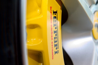 Close-up of a yellow brake caliper with the text 'Ferrari' and 'brembo carbon ceramic' printed on it. The caliper is part of a vehicle's braking system and is surrounded by the shiny surface of a wheel.