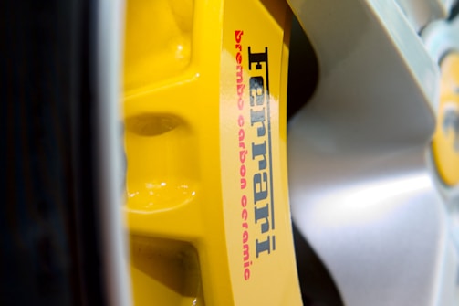 Close-up of a yellow brake caliper with the text 'Ferrari' and 'brembo carbon ceramic' printed on it. The caliper is part of a vehicle's braking system and is surrounded by the shiny surface of a wheel.