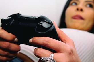 A close-up view of a person holding a video game controller with both hands. The focus is on the controller and the hands, with a ring visible on one finger. The person appears to be relaxed, resting against a pillow or cushion, with their face partially visible in the background.
