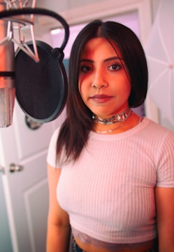 A woman with dark hair is standing in a recording studio. She is wearing a light-colored ribbed shirt and a metallic choker necklace. A large professional microphone with a pop filter is positioned in front of her. The background shows a partially open door and a studio light umbrella.