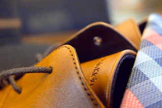 A close-up of a brown leather shoe with visible stitching, featuring a shoelace threaded through an eyelet. To the right, part of a checkered fabric pattern in blue, white, and red is visible, suggesting formal or business attire.