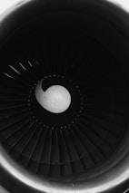A close-up view of an aircraft engine turbine, showcasing its circular pattern of blades around a central hub. The image is in black and white, highlighting the geometric and mechanical features with shadows and light contrasts.