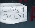 A wrinkled piece of white paper with the words 'CASH ONLY!' written in black marker is taped to a dark surface. Part of a red letter is visible in the background.