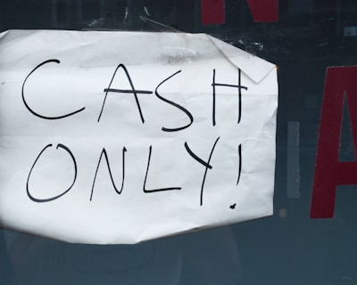 A wrinkled piece of white paper with the words 'CASH ONLY!' written in black marker is taped to a dark surface. Part of a red letter is visible in the background.