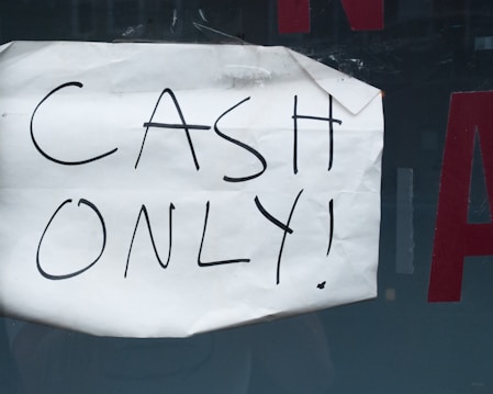 A wrinkled piece of white paper with the words 'CASH ONLY!' written in black marker is taped to a dark surface. Part of a red letter is visible in the background.