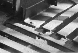 Metal beams with a worn and rugged texture are arranged in a grid pattern. Light and shadow create a stark contrast on the surfaces, highlighting the industrial nature of the scene.