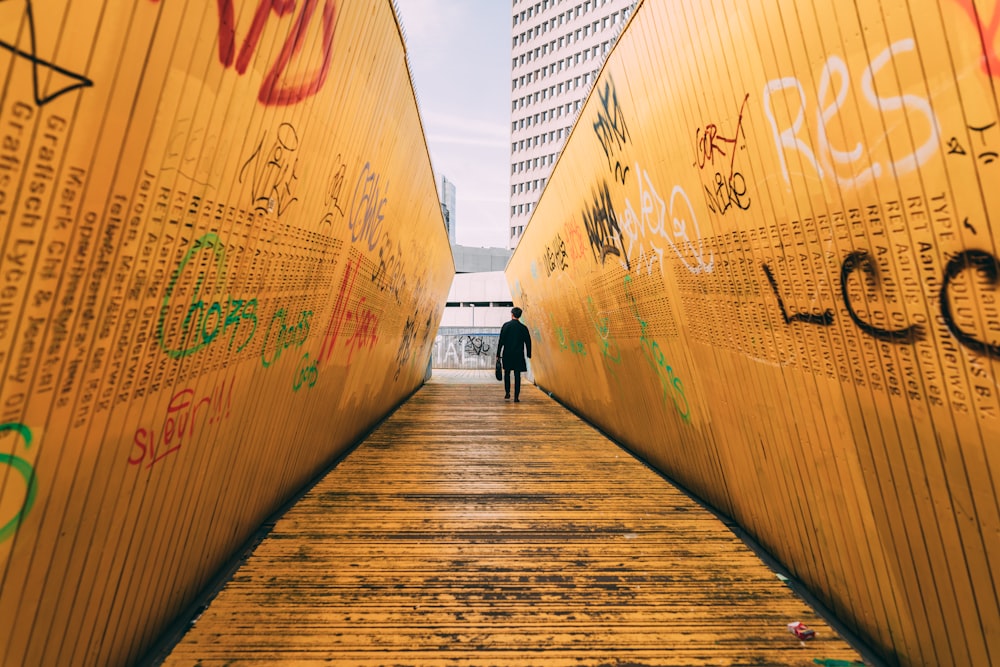 person standing on pathway between yellow wall with graffiti