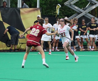 Two lacrosse players are actively engaged in a game on a green field. One player in a white Maryland uniform is moving quickly, holding a lacrosse stick, while being closely approached by a player in a red uniform wearing number 22. In the background, other team members stand on the sidelines alongside some equipment and spectators.