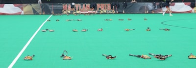A sports field with numerous lacrosse sticks arranged on the green surface. In the background, a banner displays the words 'Maryland Pride' in bold lettering. A person in sports attire walks past the sticks on the right side of the image.