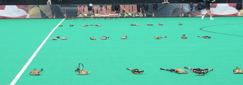 A sports field with numerous lacrosse sticks arranged on the green surface. In the background, a banner displays the words 'Maryland Pride' in bold lettering. A person in sports attire walks past the sticks on the right side of the image.