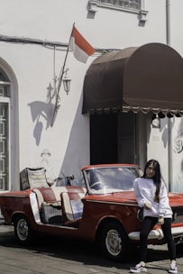 A young woman in a white long-sleeve shirt and black pants stands casually in front of a vintage red convertible car with open sides. The car is parked next to a building with white walls, adorned with an Indonesian flag and a brown awning. There's also a bicycle leaning against the wall.