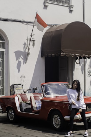 A young woman in a white long-sleeve shirt and black pants stands casually in front of a vintage red convertible car with open sides. The car is parked next to a building with white walls, adorned with an Indonesian flag and a brown awning. There's also a bicycle leaning against the wall.